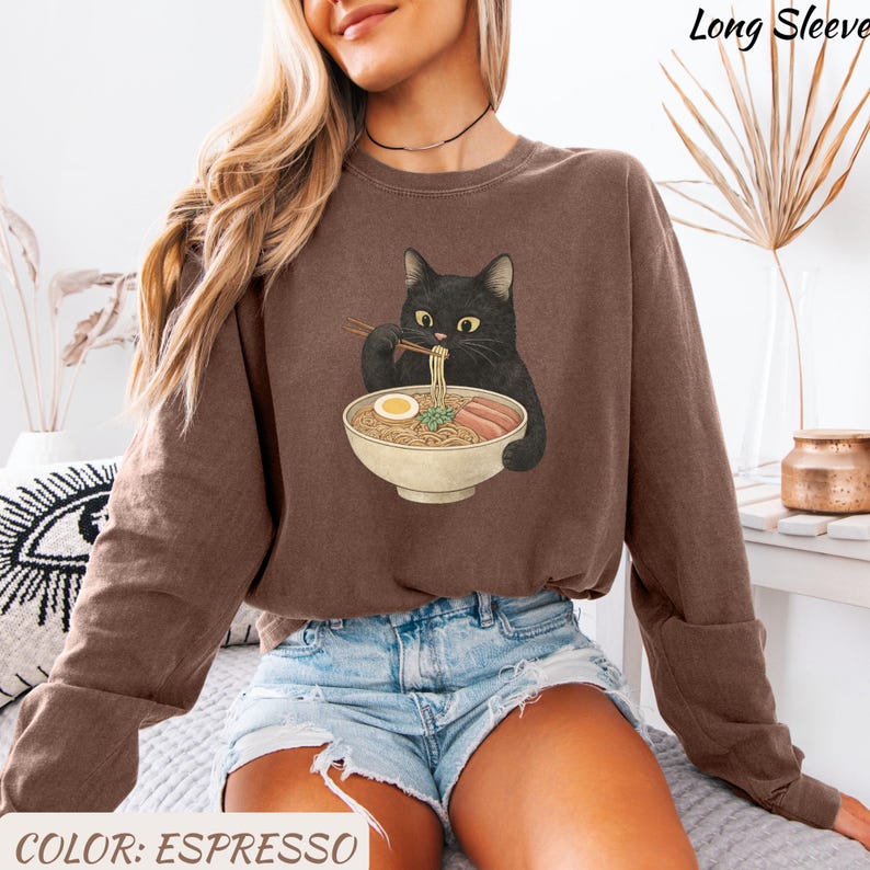 May include: Espresso-colored long-sleeve sweatshirt featuring a black cat eating ramen from a bowl. The cat is holding chopsticks. The text "Long Sleeve" is at the top right. The text "COLOR: ESPRESSO" is at the bottom.