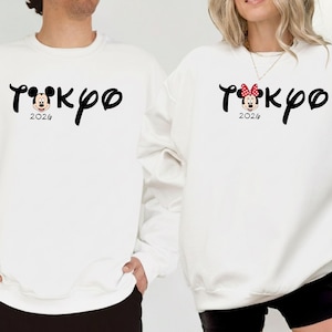 May include: White sweatshirts with the word "Tokyo" in a stylized font, featuring Mickey Mouse and Minnie Mouse designs. The year "2026" is printed below. One sweatshirt has a Mickey Mouse design, and the other has a Minnie Mouse design with a red bow.