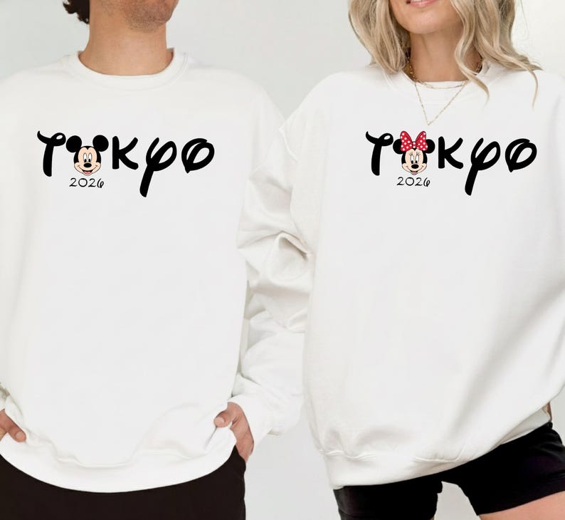 May include: White sweatshirts with the word "Tokyo" in a stylized font, featuring Mickey Mouse and Minnie Mouse designs. The year "2026" is printed below. One sweatshirt has a Mickey Mouse design, and the other has a Minnie Mouse design with a red bow.