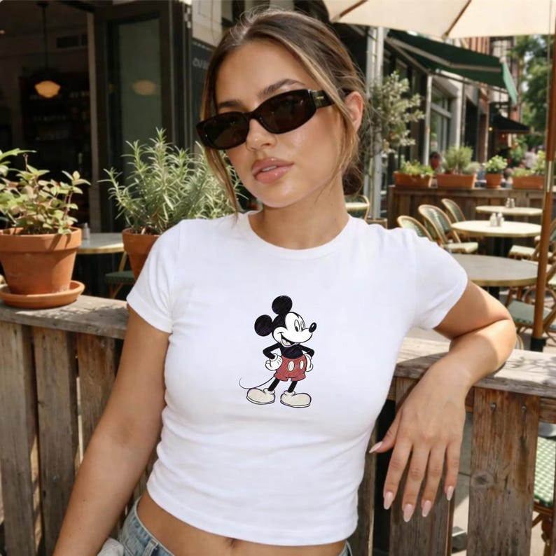 Puede incluir: Top corto blanco con un gr&aacute;fico de Mickey Mouse. El personaje de dibujos animados se muestra en su pose cl&aacute;sica, con pantalones cortos rojos y guantes blancos. La camiseta tiene mangas cortas y cuello redondo.