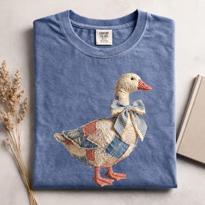 May include: A blue Comfort Colors t-shirt featuring a patchwork goose design. The goose has a cream-colored head, a red and white checkered beak, and a blue gingham bow. The body is made of various fabric patches in red, blue, and floral patterns.