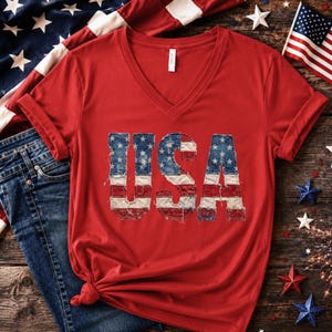 Op de afbeelding: Rood V-hals T-shirt met het woord "USA" in een distressed, patriottisch ontwerp. De letters zijn zo ontworpen dat ze op de Amerikaanse vlag lijken. Het shirt heeft opgerolde mouwen en een knoop aan de onderkant. De achtergrond omvat een spijkerbroek en de Amerikaanse vlag.