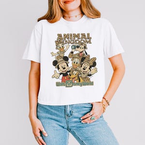 May include: White t-shirt featuring a retro-style graphic of Disney characters, including Mickey and Minnie Mouse, Donald Duck, and Goofy. The design includes the text "ANIMAL KINGDOM" and "Walt Disney World" in a vintage font.