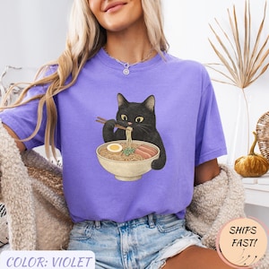 May include: Violet-colored t-shirt featuring a black cat enjoying ramen. The cat is holding chopsticks and eating noodles from a bowl. The shirt has the text "COLOR: VIOLET" at the bottom and a "SHIPS FAST!" sticker.