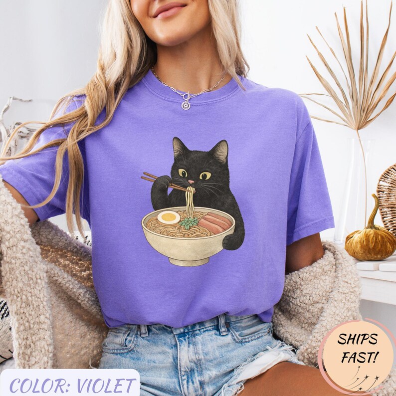 May include: Violet-colored t-shirt featuring a black cat enjoying ramen. The cat is holding chopsticks and eating noodles from a bowl. The shirt has the text "COLOR: VIOLET" at the bottom and a "SHIPS FAST!" sticker.