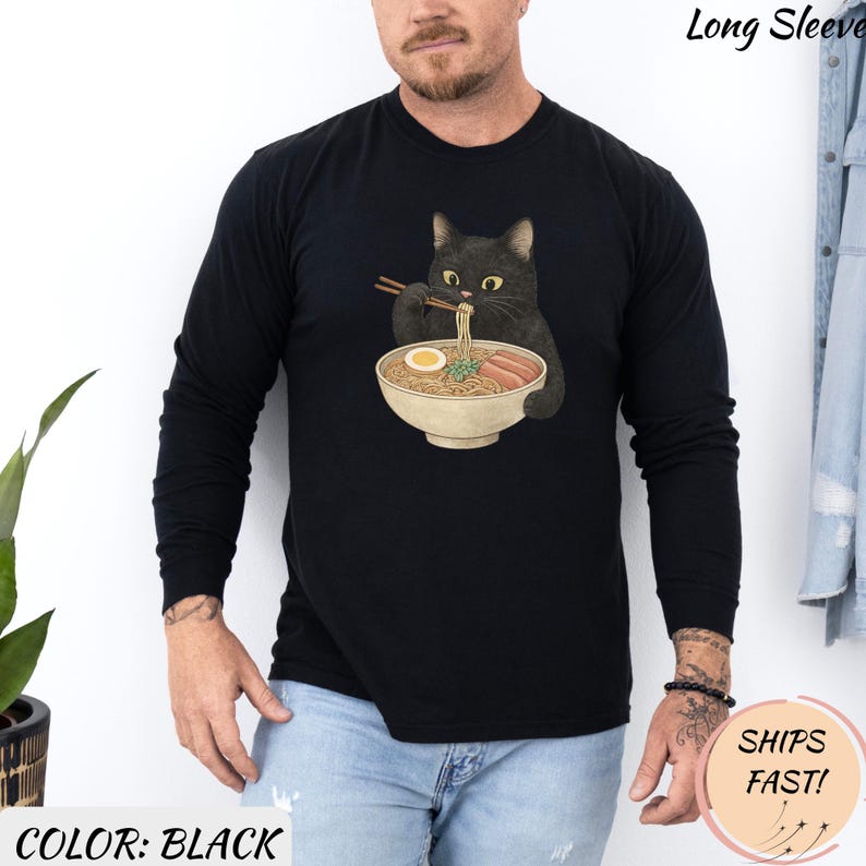 May include: Black long-sleeve shirt featuring a graphic of a black cat eating ramen from a bowl with chopsticks. The shirt has the text "Long Sleeve" at the top and "COLOR: BLACK" at the bottom. The shirt is a solid black color.