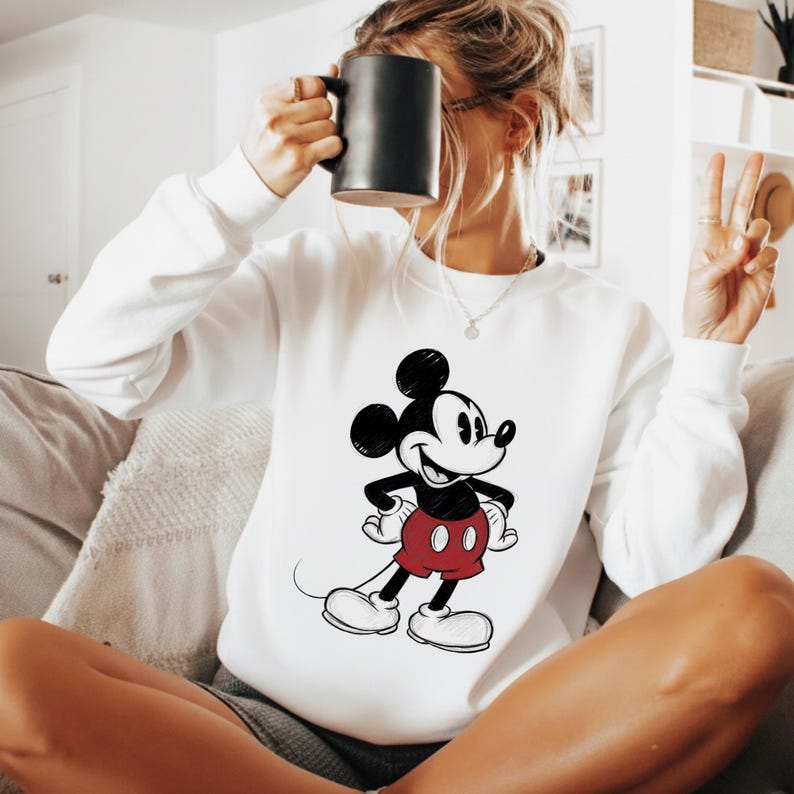 May include: White sweatshirt featuring a classic Mickey Mouse graphic. The cartoon character is depicted in black, red, and white, with a smiling expression. The sweatshirt is a casual, comfortable style.