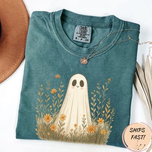 May include: Teal Comfort Colors t-shirt featuring a friendly ghost illustration surrounded by orange flowers and greenery. A delicate gold necklace rests on the shirt. The shirt has a crew neck and short sleeves. The tag reads "S - P - CH".