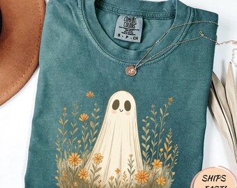 May include: Teal Comfort Colors t-shirt with a friendly ghost illustration amidst orange flowers and greenery. A delicate gold necklace rests on the shirt. The shirt has a crew neck and short sleeves. The tag reads "S - P - CH".