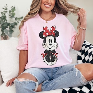 May include: A light pink t-shirt featuring a graphic of Minnie Mouse. Minnie is depicted in a seated pose, wearing her signature red and white polka dot bow and shoes. The shirt is a casual style, suitable for everyday wear.