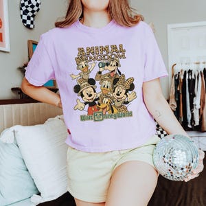 May include: A light purple t-shirt featuring a graphic of Disney characters, including Mickey and Minnie Mouse, Donald Duck, and Goofy. The text "ANIMAL KINGDOM" and "Walt Disney World" are printed on the shirt in a retro style.
