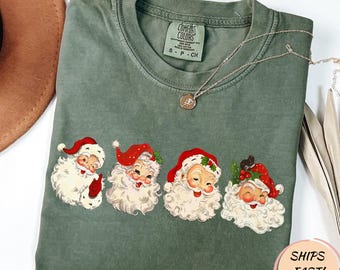 May include: An olive green Comfort Colors t-shirt featuring four vintage Santa Claus faces with red hats and white beards. A silver necklace with a pendant is draped across the shirt. The shirt has a crew neck and short sleeves.