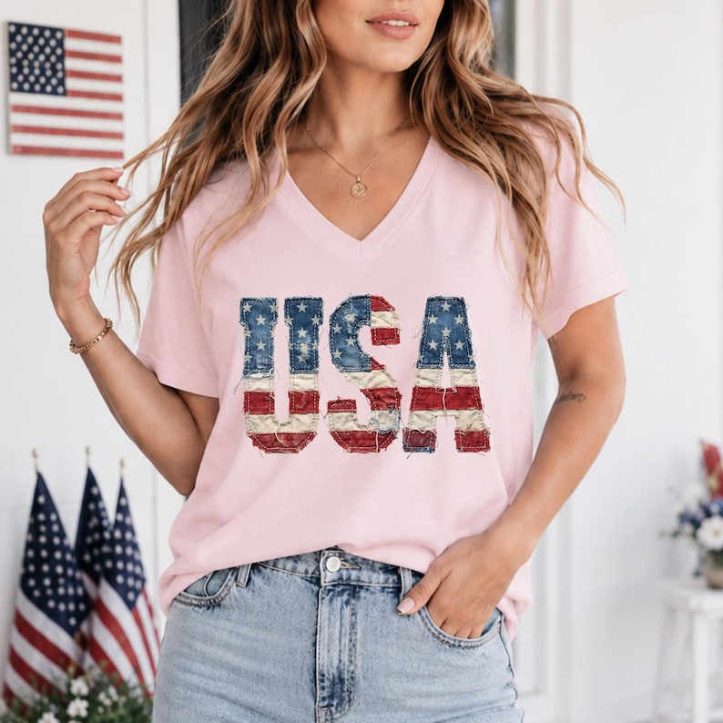 Op de afbeelding: Lichtroze V-hals T-shirt met het woord "USA" op de voorkant gedrukt in een versleten Amerikaans vlagontwerp. De letters zijn blauw met witte sterren en rood-witte strepen. Het shirt wordt gedragen met een blauwe jeans.