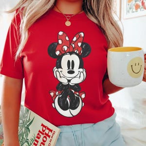 May include: Red t-shirt featuring a Minnie Mouse graphic with a red bow and white polka dots. The shirt is paired with a gold necklace and a white mug with a yellow interior and a smiley face.