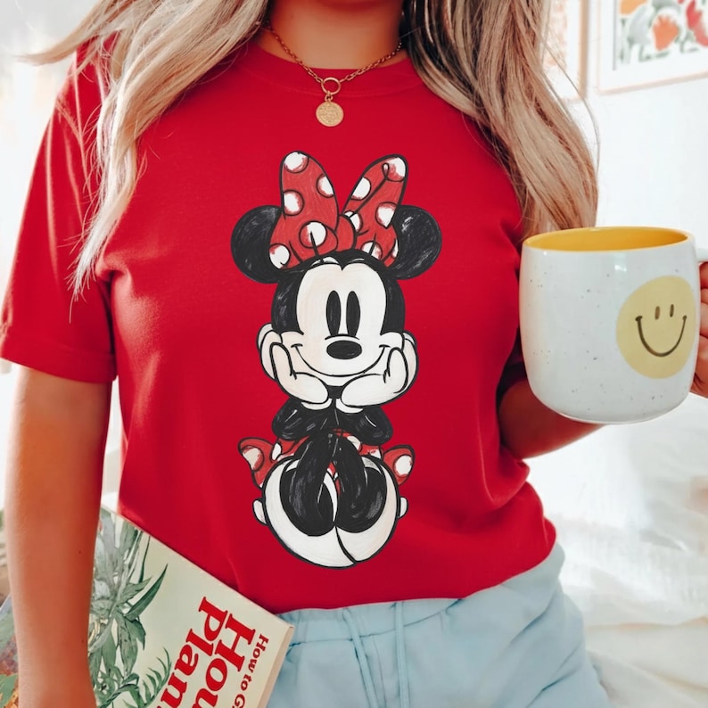 May include: Red t-shirt featuring a Minnie Mouse graphic with a red bow and white polka dots. The shirt is paired with a gold necklace and a white mug with a yellow interior and a smiley face.