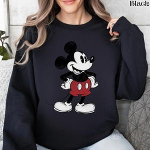 May include: Black sweatshirt featuring a classic Mickey Mouse cartoon graphic. The graphic shows Mickey Mouse in his iconic pose, wearing red shorts, white gloves, and shoes. The word "Black" is printed in the upper right corner.