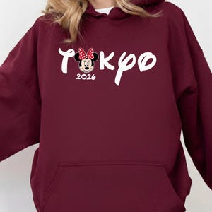 May include: Maroon hoodie with a front pocket and a white drawstring. The front features the word "Tokyo" in a script font, with a Minnie Mouse graphic and the year "2026".