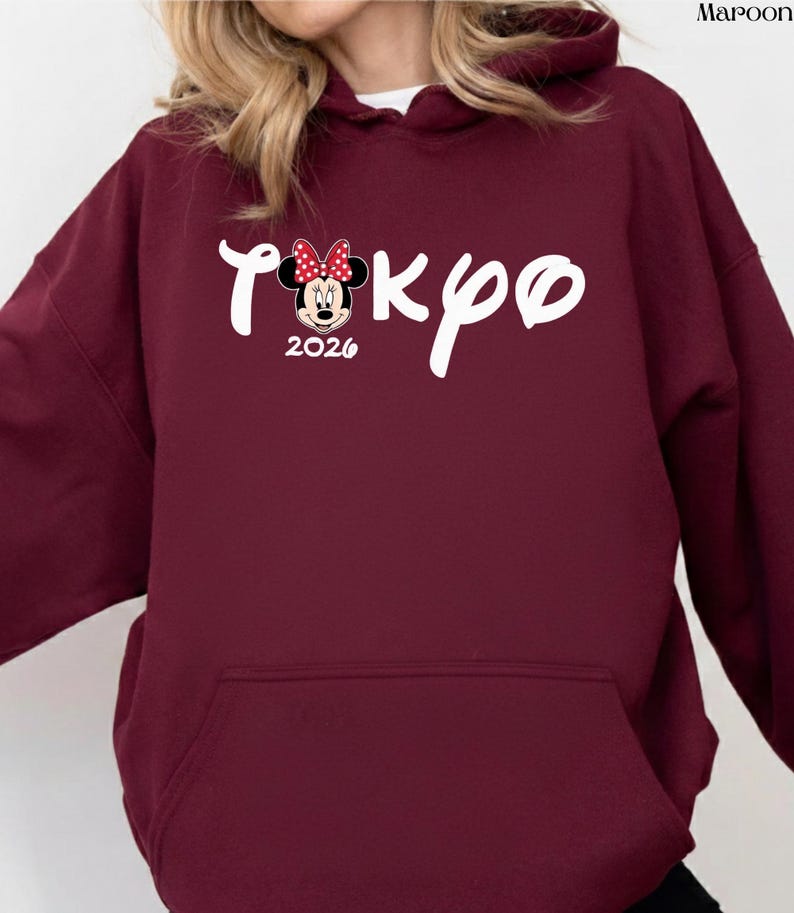 May include: Maroon hoodie with a front pocket and a white drawstring. The front features the word "Tokyo" in a script font, with a Minnie Mouse graphic and the year "2026".