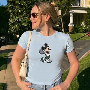 May include: Light blue t-shirt featuring a cartoon Mickey Mouse playing tennis. The graphic shows Mickey in a green and white tennis outfit, holding a racket. The shirt has short sleeves and a crew neck.