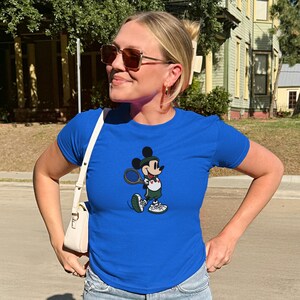 May include: A royal blue t-shirt featuring a cartoon Mickey Mouse playing tennis. Mickey is wearing a green and white tennis outfit and holding a tennis racket. The shirt has short sleeves and a crew neck.