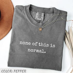 Peut inclure: T-shirt Comfort Colors de couleur poivre avec l'inscription blanche « none of this is normal ». Un collier argenté avec un pendentif est posé sur le t-shirt. Le t-shirt est plié sur une surface blanche.