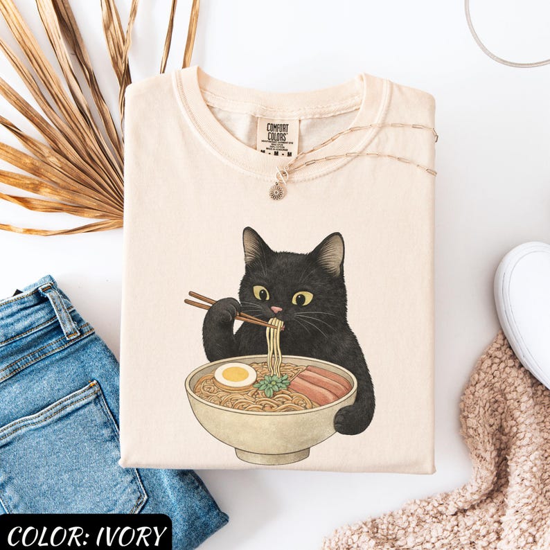 May include: Ivory-colored t-shirt featuring a black cat eating ramen from a bowl. The cat is holding chopsticks and the bowl contains noodles, egg, and meat. The shirt has the text "COMFORT COLORS" on the tag. The color of the shirt is ivory.