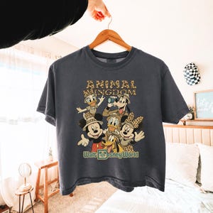 May include: A dark gray t-shirt featuring a graphic of Disney characters in safari attire. The text "ANIMAL KINGDOM" is above the characters, and "Walt Disney World" is below. The shirt is hanging on a wooden hanger.