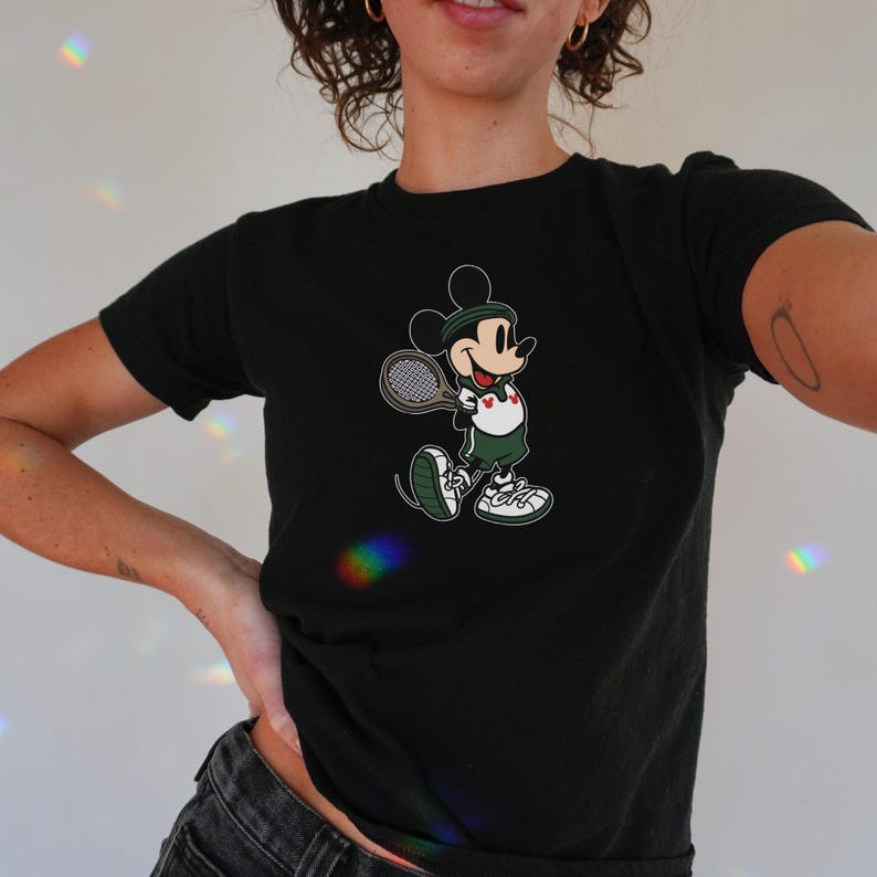 May include: Black t-shirt featuring a cartoon Mickey Mouse playing tennis. Mickey is wearing a green tennis outfit, a white shirt with a heart, and white sneakers. He is holding a tennis racket. The design is centered on the shirt.