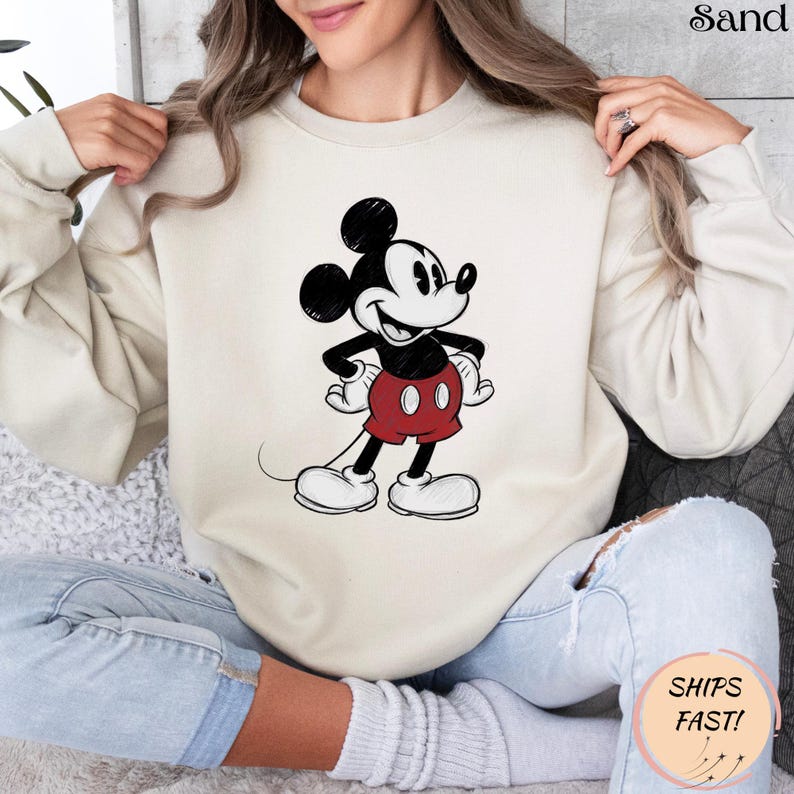 May include: Sand-colored sweatshirt featuring a classic Mickey Mouse graphic. The cartoon character is depicted in black, white, and red, with his signature shorts and gloves. The sweatshirt has a crew neck and long sleeves, offering a casual and comfortable style.