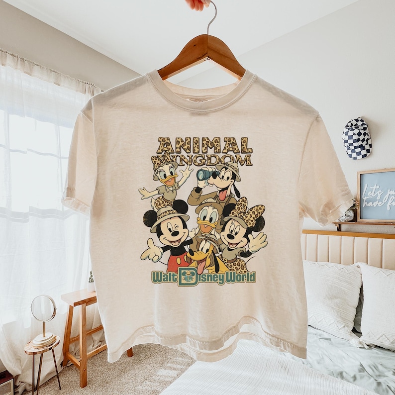 May include: Cream-colored t-shirt featuring a graphic of Disney characters, including Mickey and Minnie Mouse, Donald and Daisy Duck, and Goofy. The text "ANIMAL KINGDOM" and "Walt Disney World" are printed on the shirt. The shirt is hanging on a wooden hanger.