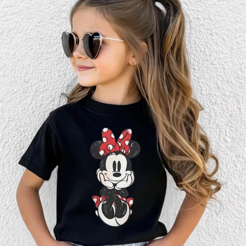 May include: Black t-shirt featuring a Minnie Mouse graphic. The graphic shows Minnie Mouse with a red polka dot bow and red shoes. The shirt is a crew neck style.
