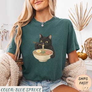 May include: Blue spruce t-shirt featuring a black cat eating ramen from a bowl with chopsticks. The graphic includes noodles, egg, and meat. The text "COLOR: BLUE SPRUCE" is at the bottom. The shirt is a relaxed fit.