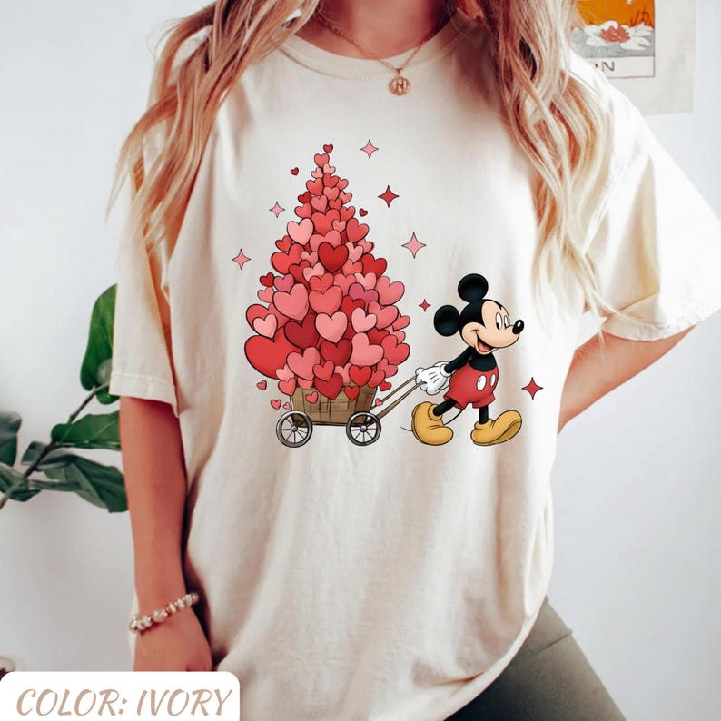 Puede incluir: Camiseta color marfil con un dise&ntilde;o de Mickey Mouse tirando de un carro lleno de decoraciones en forma de coraz&oacute;n. El dise&ntilde;o incluye un &aacute;rbol hecho de corazones y acentos de estrellas rojas, ideal para San Valent&iacute;n. El texto "COLOR: IVORY" est&aacute; en la parte inferior.