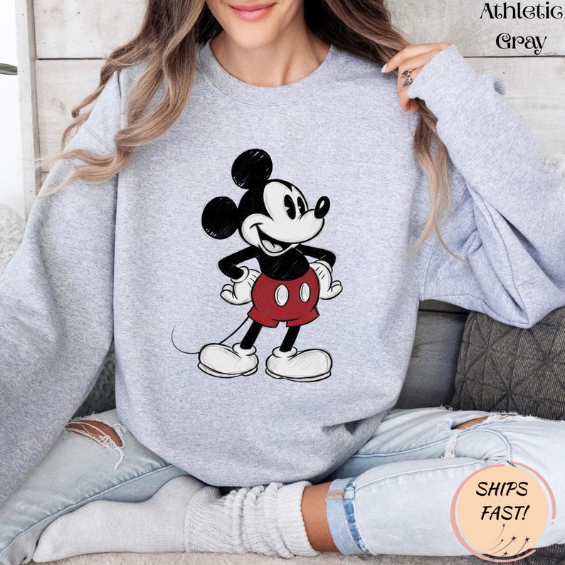May include: Athletic gray sweatshirt featuring a classic Mickey Mouse graphic. The cartoon character is depicted in black, white, and red, with his signature shorts and gloves. The sweatshirt has long sleeves and a crew neck.