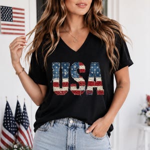 Op de afbeelding: Zwart V-hals T-shirt met het woord "USA" in een versleten, patriottisch ontwerp. De letters hebben een denim-achtige textuur met de sterren en strepen van de Amerikaanse vlag. Het shirt wordt gedragen met een lichtblauwe spijkerbroek.