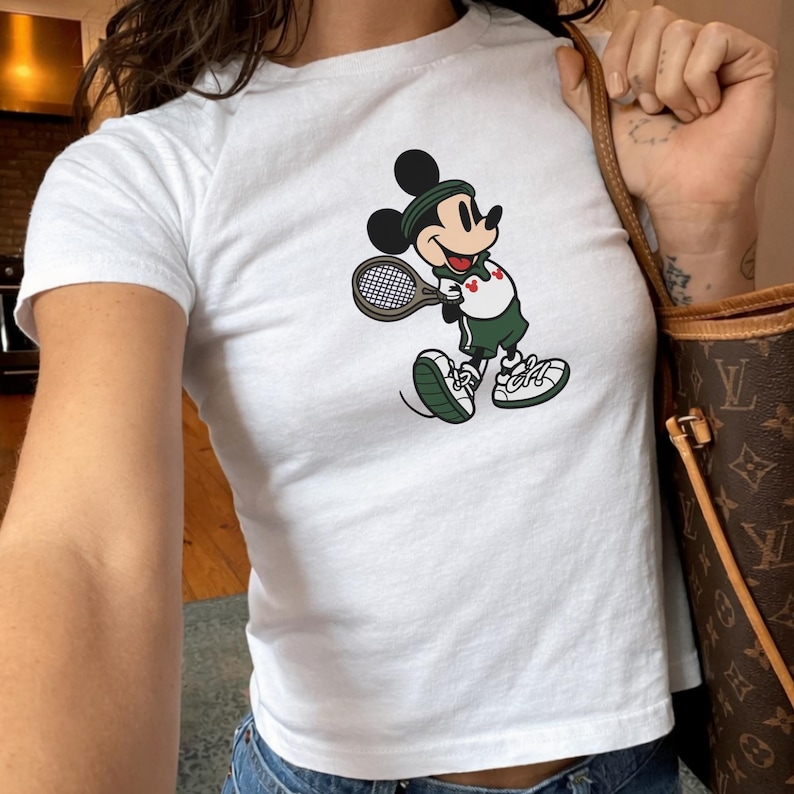 May include: White t-shirt featuring a cartoon Mickey Mouse playing tennis. Mickey is wearing a green headband, shorts, and shoes, holding a tennis racket. The shirt is short-sleeved and has a crew neck.