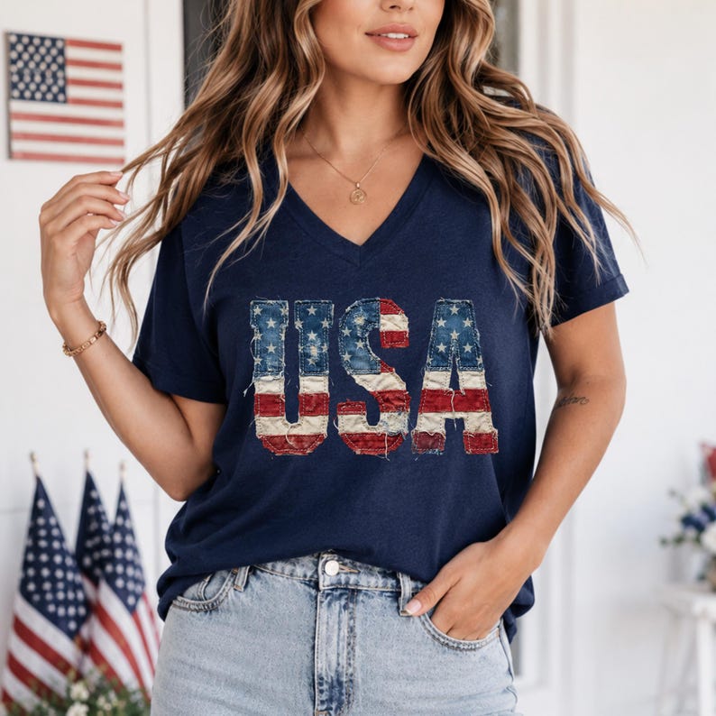 Op de afbeelding: Marineblauwe V-hals T-shirt met het woord "USA" in een versleten Amerikaans vlagontwerp. Het shirt wordt gecombineerd met lichtblauwe denim jeans. De achtergrond toont Amerikaanse vlaggen, wat duidt op een patriottisch thema.
