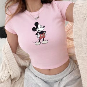Puede incluir: Un top corto rosa claro con un dise&ntilde;o cl&aacute;sico de Mickey Mouse. La camiseta de manga corta tiene cuello redondo y est&aacute; hecha de un material suave. El personaje de dibujos animados se representa en su pose ic&oacute;nica, con pantalones cortos rojos y guantes blancos.