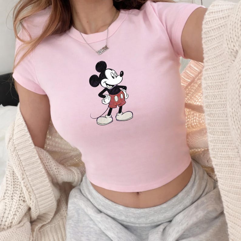 Puede incluir: Un top corto rosa claro con un dise&ntilde;o cl&aacute;sico de Mickey Mouse. La camiseta de manga corta tiene cuello redondo y est&aacute; hecha de un material suave. El personaje de dibujos animados se representa en su pose ic&oacute;nica, con pantalones cortos rojos y guantes blancos.