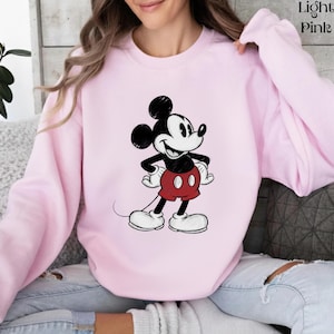 May include: Light pink sweatshirt featuring a classic Mickey Mouse graphic. The cartoon character is depicted in black, white, and red, with his signature large ears and smiling face. The sweatshirt is a solid light pink color.