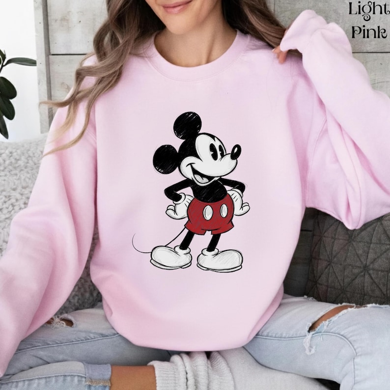 May include: Light pink sweatshirt featuring a classic Mickey Mouse graphic. The cartoon character is depicted in black, white, and red, with his signature large ears and smiling face. The sweatshirt is a solid light pink color.
