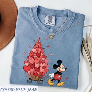 Puede incluir: Camiseta azul vaquero con un gr&aacute;fico de Mickey Mouse tirando de un carro lleno de globos en forma de coraz&oacute;n. La camiseta tiene cuello redondo y mangas cortas. Tambi&eacute;n se ve un collar dorado con un colgante.