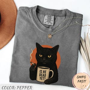Może przedstawiać: Koszulka Comfort Colors w kolorze pieprzu z ilustracją czarnego kota z papierosem i kubkiem z napisem "IS HE DEAD YET?". Kot jest umieszczony na pomarańczowym okręgu. Okrągły dekolt i kr&oacute;tkie rękawy.