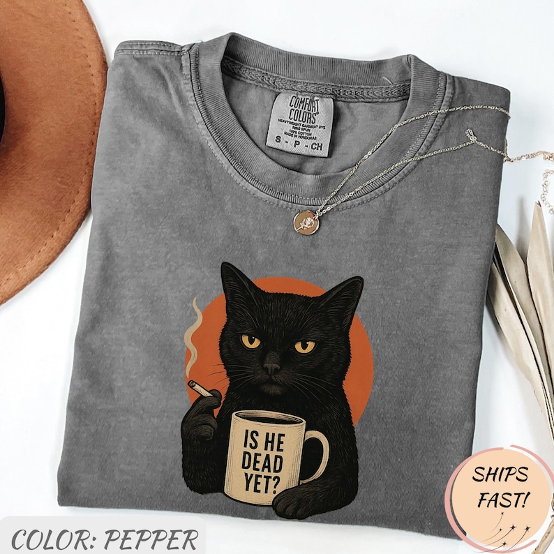 Może przedstawiać: Koszulka Comfort Colors w kolorze pieprzu z ilustracją czarnego kota z papierosem i kubkiem z napisem "IS HE DEAD YET?". Kot jest umieszczony na pomarańczowym okręgu. Okrągły dekolt i kr&oacute;tkie rękawy.