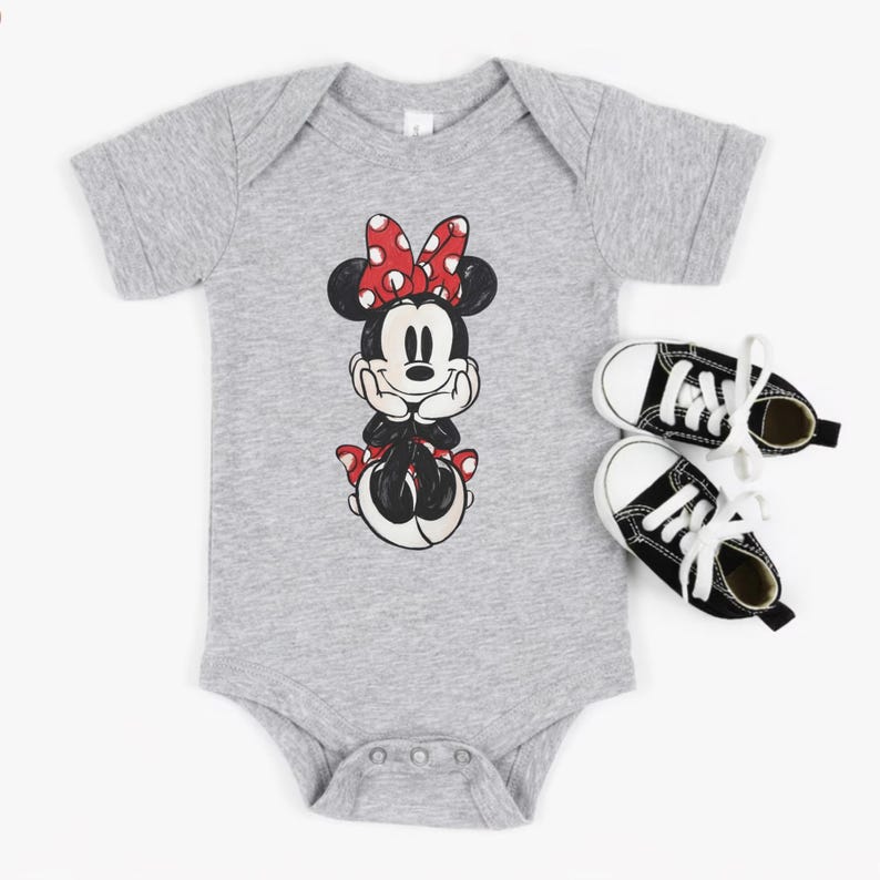 May include: A gray baby onesie featuring a Minnie Mouse graphic with a red bow and black and white shoes. The onesie has a snap closure at the bottom. The shoes are black with white laces and soles.