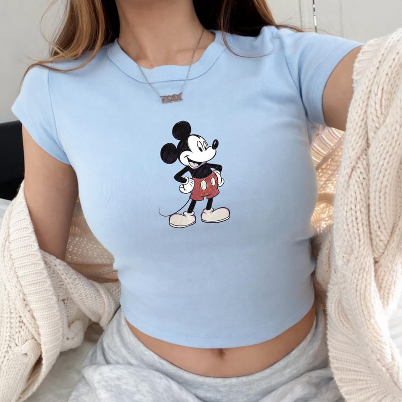 Puede incluir: Top corto azul claro con un estampado de Mickey Mouse. La camiseta de manga corta tiene cuello redondo y est&aacute; hecha de un tejido suave. El personaje de dibujos animados es de color negro, rojo y blanco.