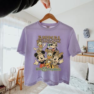 May include: A light purple t-shirt featuring a graphic of Disney characters, including Mickey Mouse, Minnie Mouse, Donald Duck, Daisy Duck, Goofy, and Pluto. The shirt has the words "ANIMAL KINGDOM" and "Walt Disney World" printed on it.