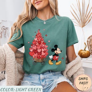 Puede incluir: Camiseta verde claro con un dise&ntilde;o de Mickey Mouse tirando de un carro lleno de decoraciones en forma de coraz&oacute;n. El texto "COLOR: LIGHT GREEN" est&aacute; en la parte inferior. El dise&ntilde;o incluye corazones y estrellas rosas.