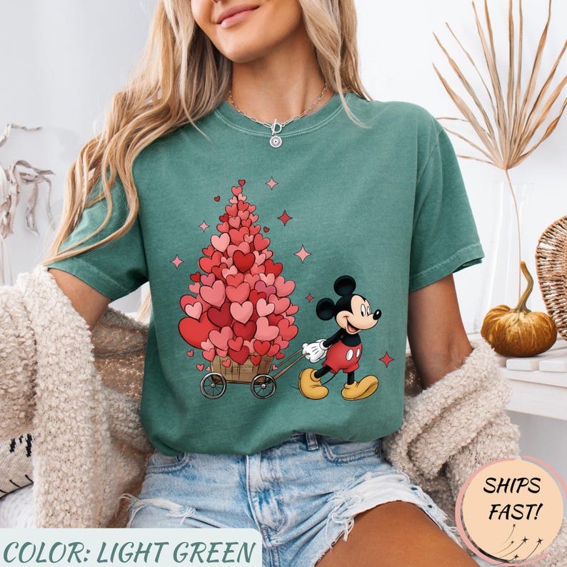 Puede incluir: Camiseta verde claro con un dise&ntilde;o de Mickey Mouse tirando de un carro lleno de decoraciones en forma de coraz&oacute;n. El texto "COLOR: LIGHT GREEN" est&aacute; en la parte inferior. El dise&ntilde;o incluye corazones y estrellas rosas.