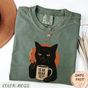 Może przedstawiać: Koszulka Comfort Colors w kolorze mchu z czarnym kotem trzymającym papierosa i kubek z napisem "IS HE DEAD YET?". Kot jest umieszczony na pomarańczowym okręgu. Srebrny naszyjnik jest na koszulce.